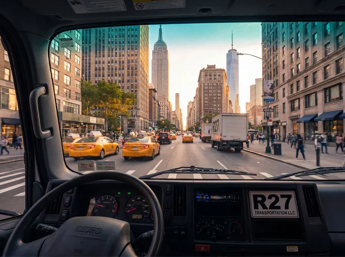 The view from inside an R27 Transportation truck cab driving up a New York City avenue toward the Empire State Building and One World Trade Center.